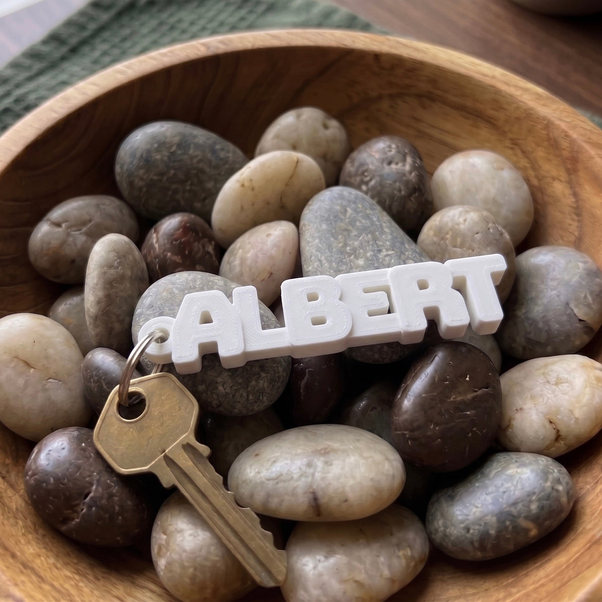 White custom name keychain reading ALBERT with a key in a bowl of pebbles