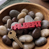 Red custom name keychain reading ALBERT with a key in a bowl of pebbles