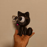 Swag Layers black 3D printed cat glasses holder with gold-rimmed spectacles held in a hand against a plain wall