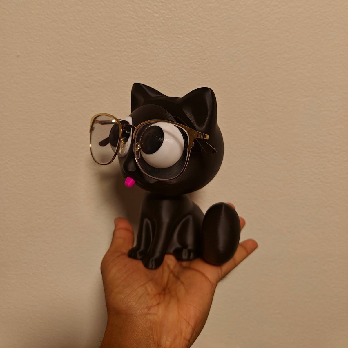 Swag Layers black 3D printed cat glasses holder with gold-rimmed spectacles held in a hand against a plain wall