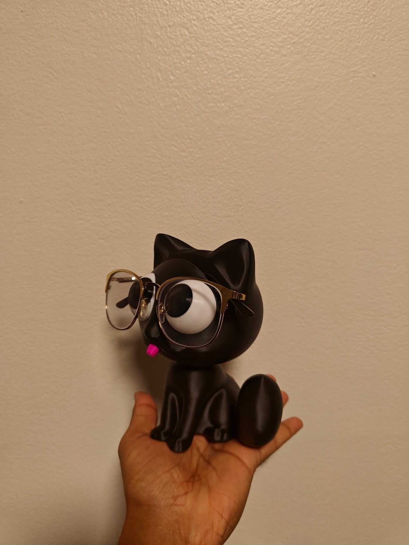 Swag Layers black 3D printed cat glasses holder with gold-rimmed spectacles held in a hand against a plain wall