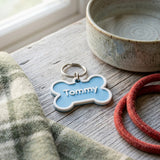 Swag Layers personalized blue and white 3D-printed bone-shaped dog tag with the name Tommy on a rustic wooden surface.